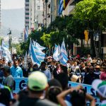 A lively urban protest with people holding flags and signs on a sunny day.